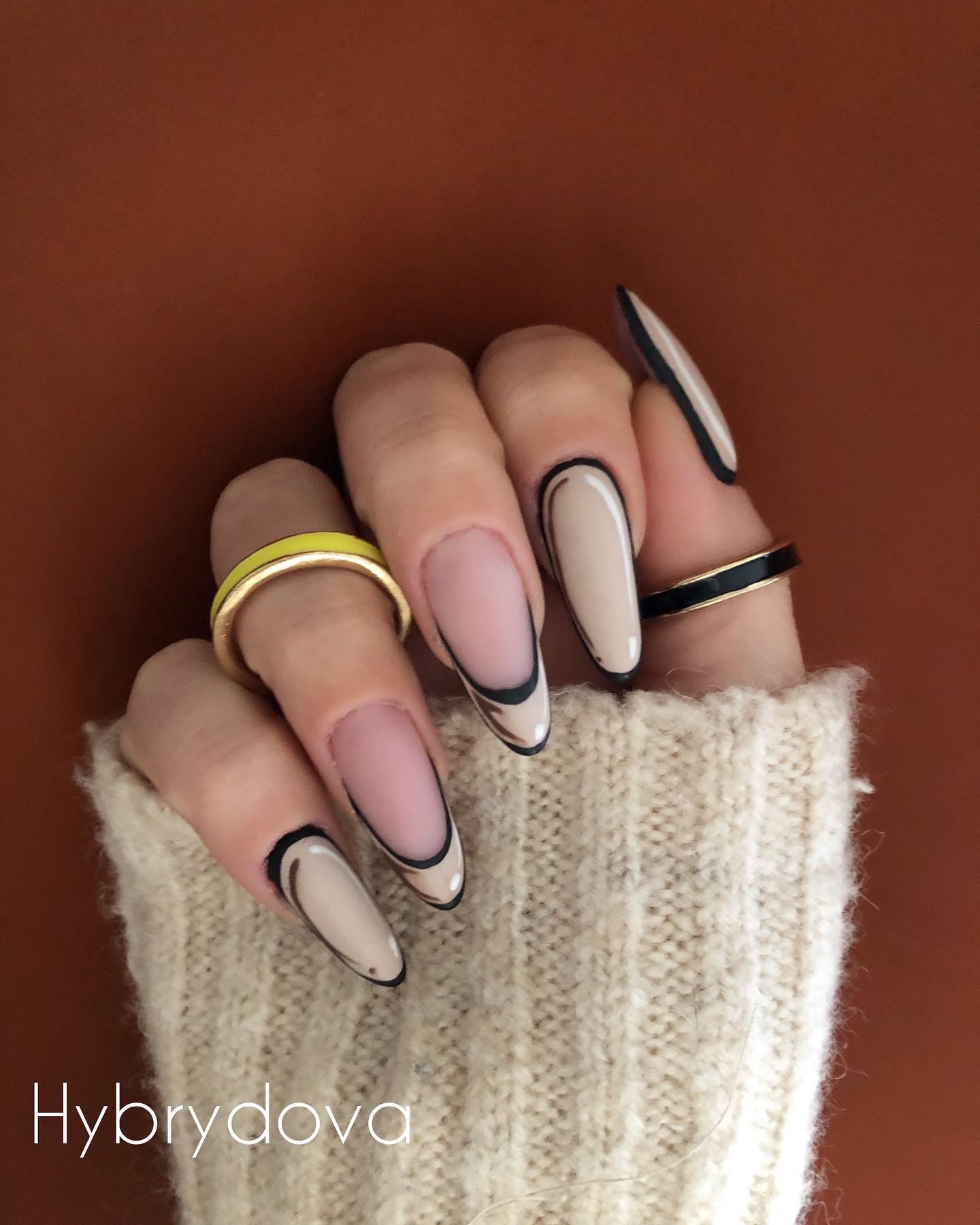 Dłoń w swetrze z beżowymi paznokciami w stylu kreskówkowym, z czarnym obrysem i pierścionkami w kolorze żółtym i czarnym.