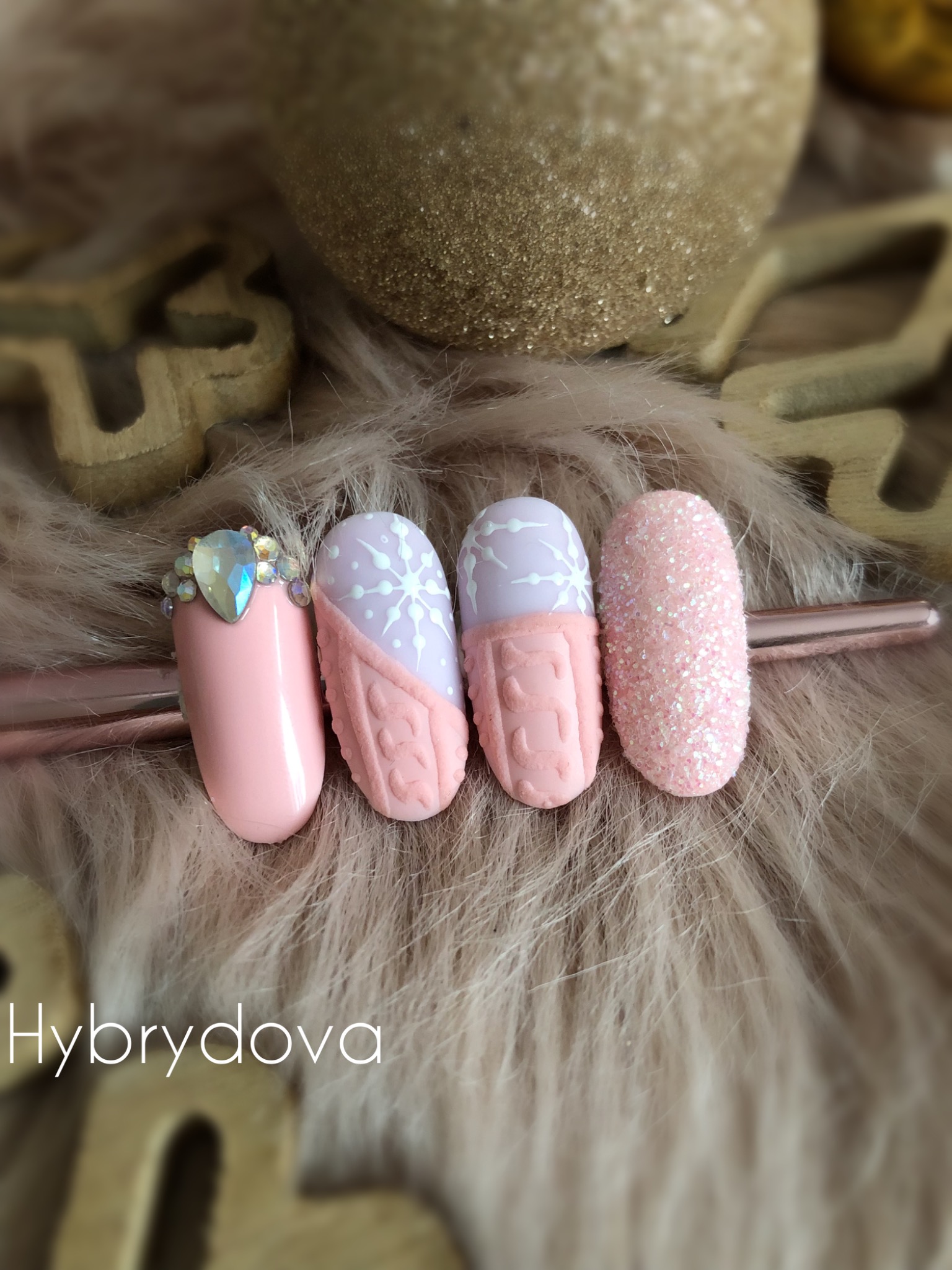 Cztery pastelowe paznokcie z zimowymi zdobieniami: różowy z kryształkami, liliowy ze śnieżynką i wzorem sweterkowym, różowy ze wzorem sweterkowym, różowy brokatowy, na futrzanym tle z dekoracjami.