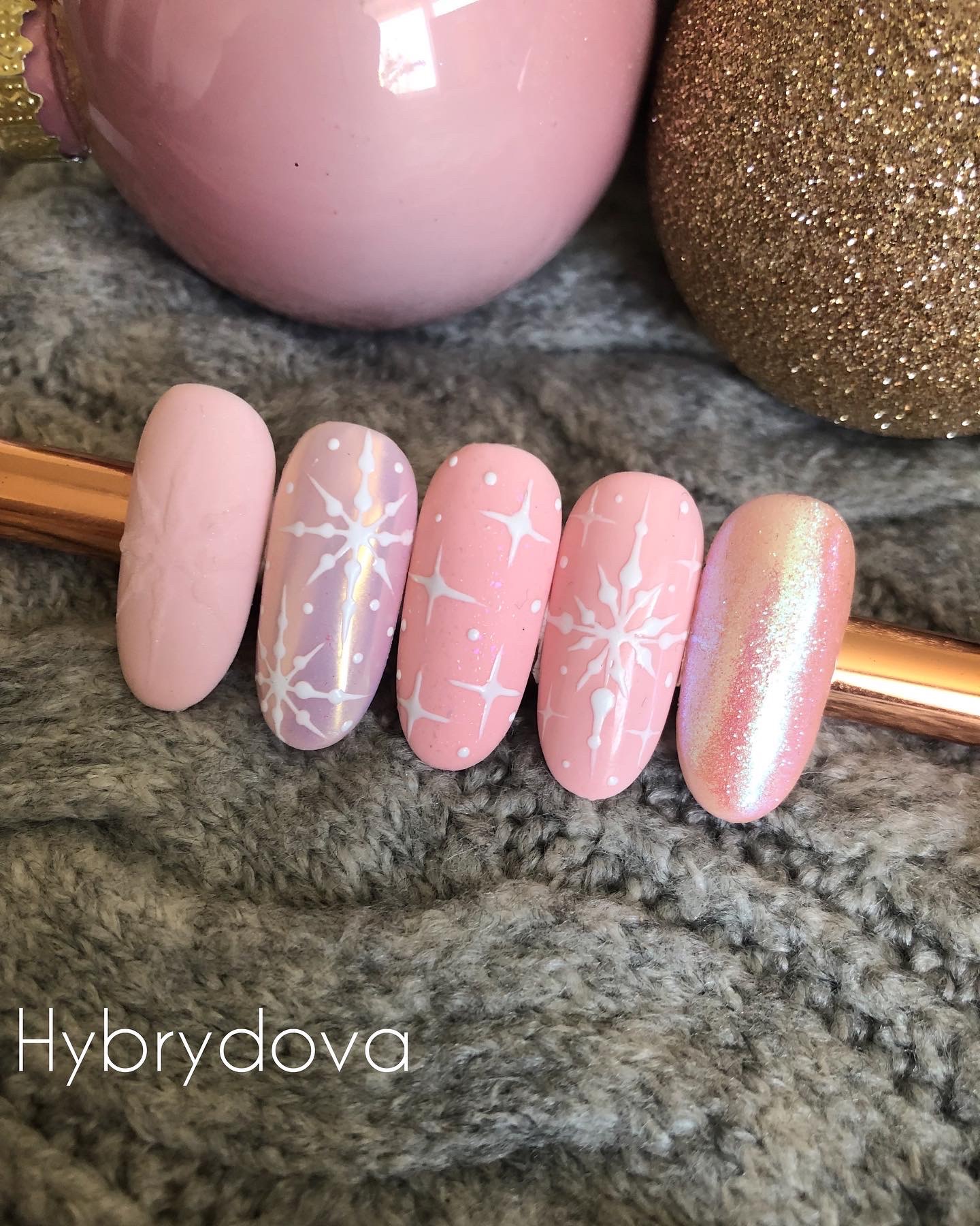 Różowe paznokcie hybrydowe z zimowymi wzorami śnieżynek, ułożone na szarym, wełnianym tle z dekoracjami świątecznymi.