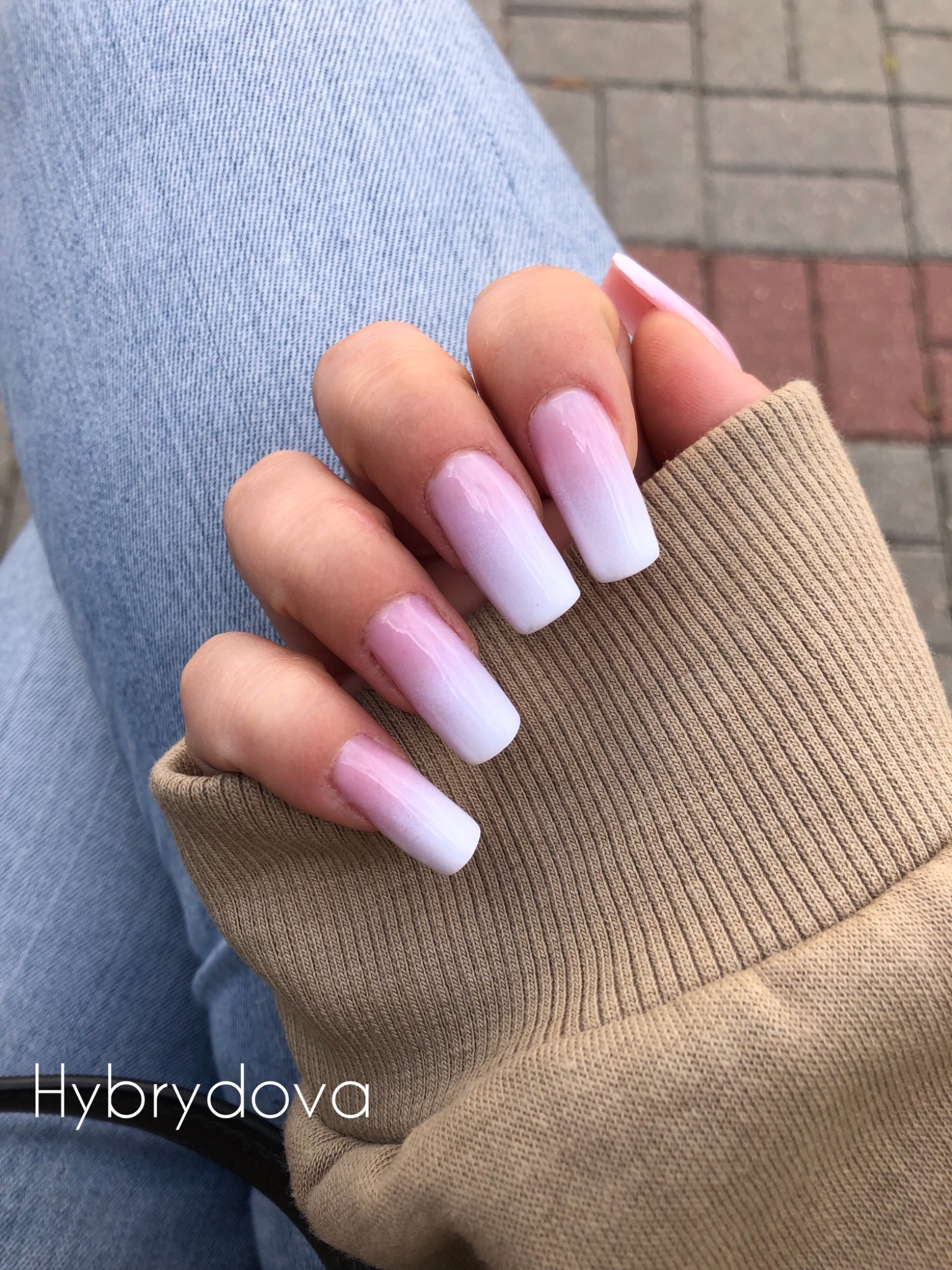 Dłoń z kwadratowymi paznokciami ombre, przechodzącymi od różu u nasady do bieli na końcach, na tle dżinsów i beżowej bluzy.