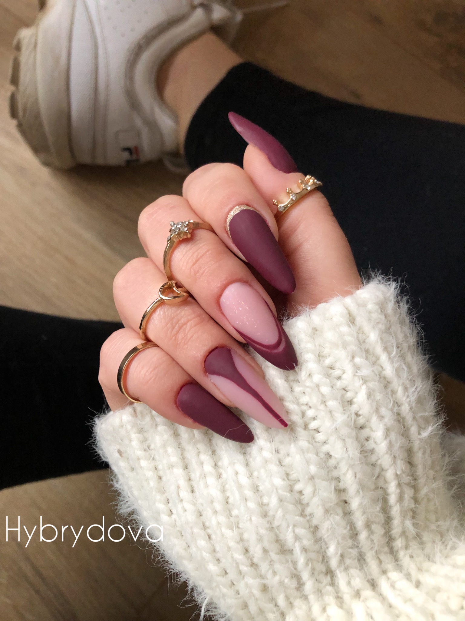 Dłoń z burgundowymi, matowymi paznokciami w różnych stylizacjach, zdobiona złotymi pierścionkami, na tle białego swetra i buta sportowego.