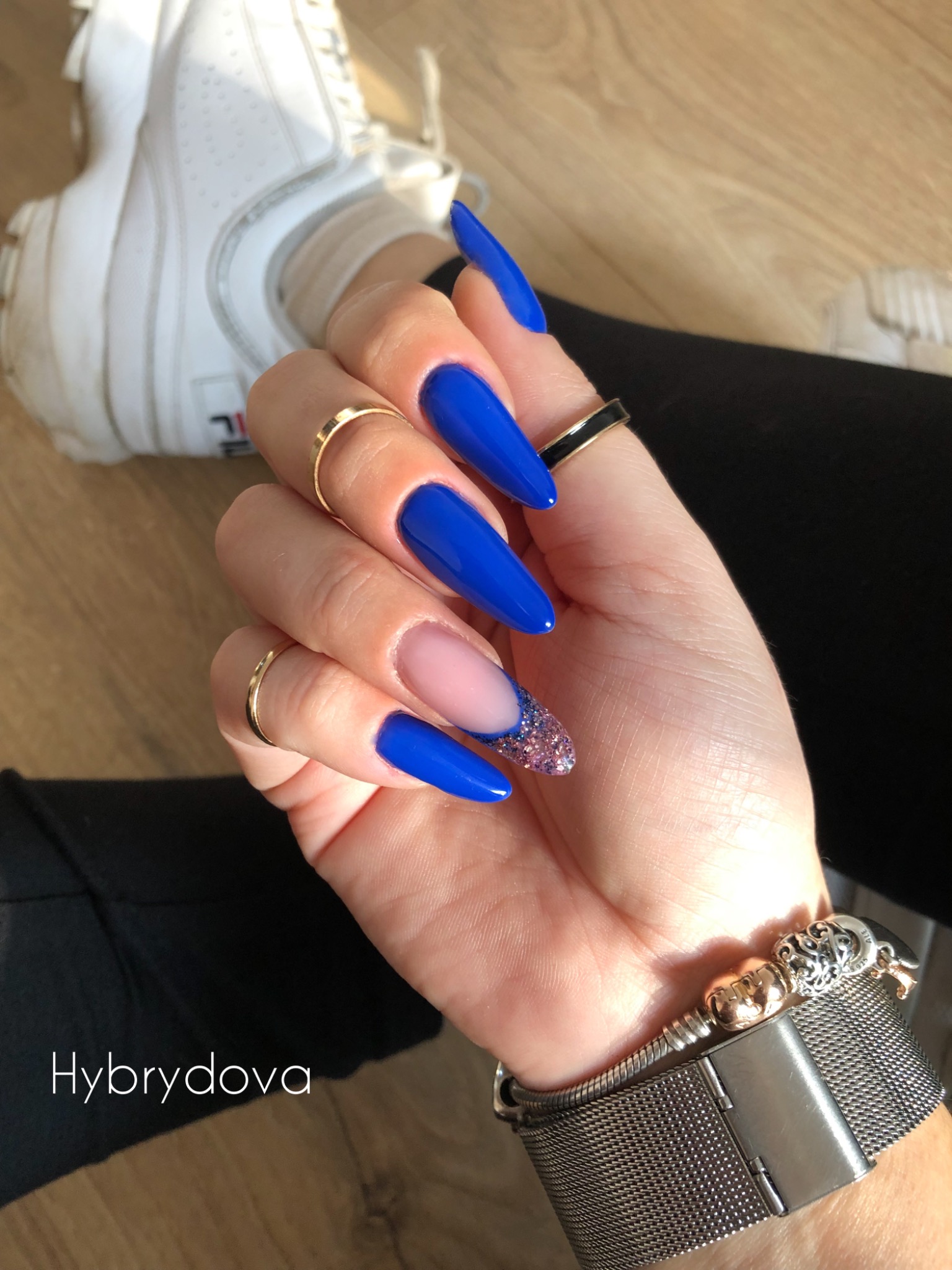 Dłoń z długimi, kobaltowymi paznokciami w kształcie migdała, jeden paznokieć z brokatowym zdobieniem french, złote pierścionki na palcach, srebrna bransoletka Pandora, białe sneakersy w tle.