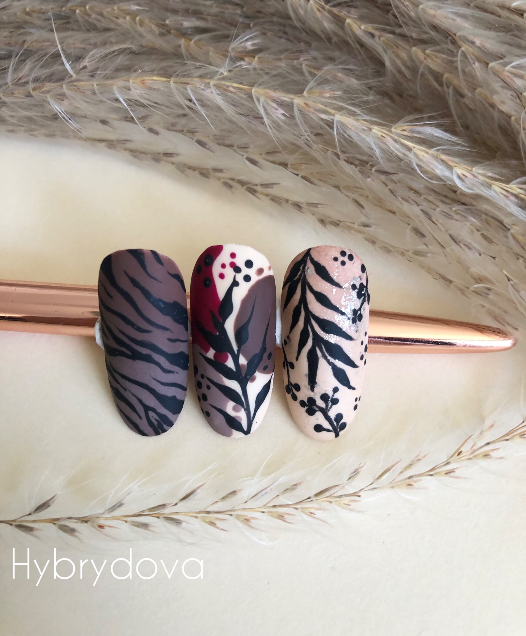 Trzy pomalowane paznokcie z różnymi wzorami (tygrysi, liście, abstrakcja) prezentowane na tle z suszonych traw, wykonane w Rypinie.