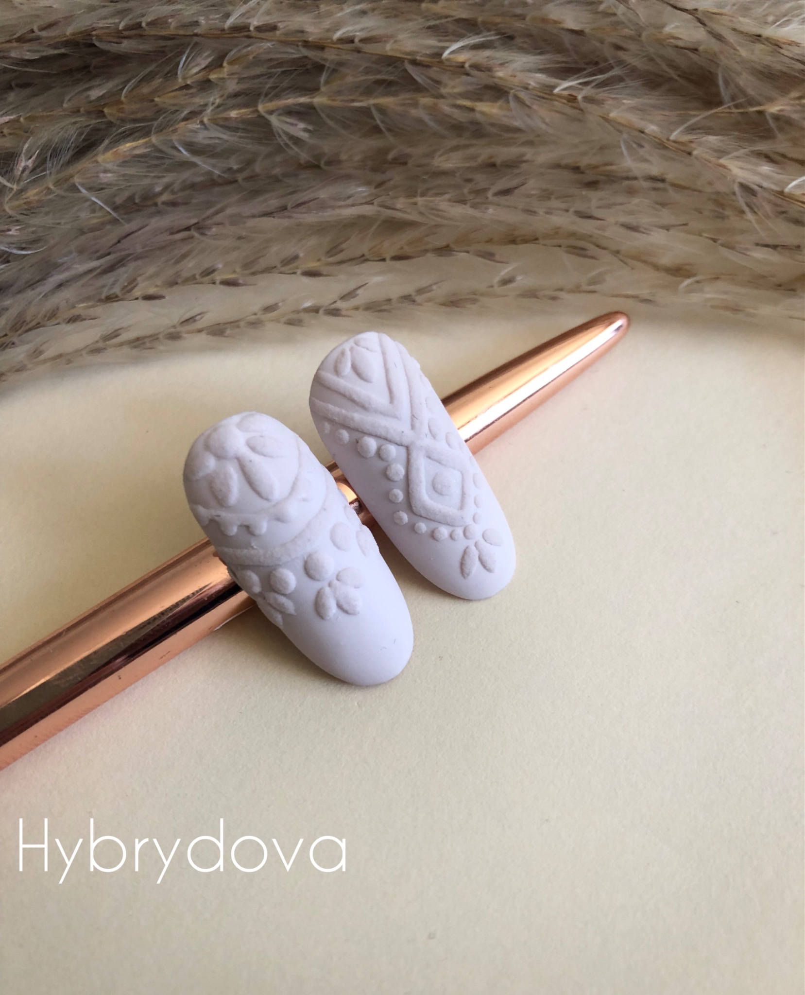 Dwa białe tipsy z wypukłymi, etnicznymi wzorami leżące na miedzianym długopisie, na tle beżowego papieru i puszystej trawy.