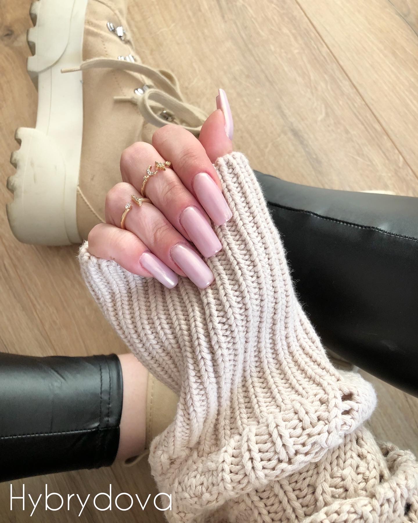 Dłoń w kremowym swetrze prezentuje długie, kwadratowe paznokcie w odcieniu pudrowego różu, ozdobione delikatnymi, złotymi pierścionkami na tle drewnianej podłogi, beżowych butów i czarnych...