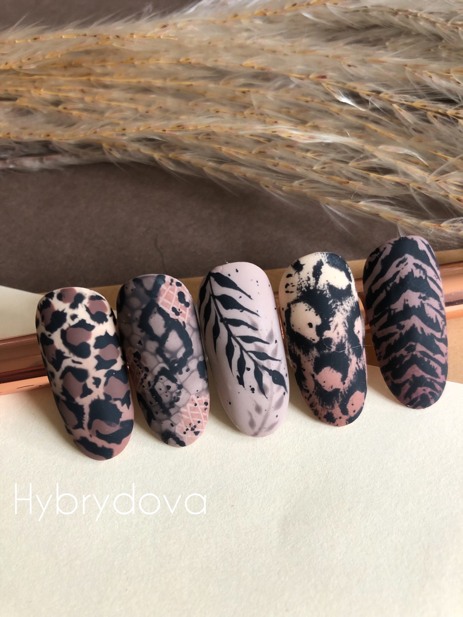 Pięć matowych paznokci z różnymi wzorami zwierzęcymi (lampart, wąż, zebra, tygrys) i liśćmi na beżowym tle z suchymi trawami w tle.