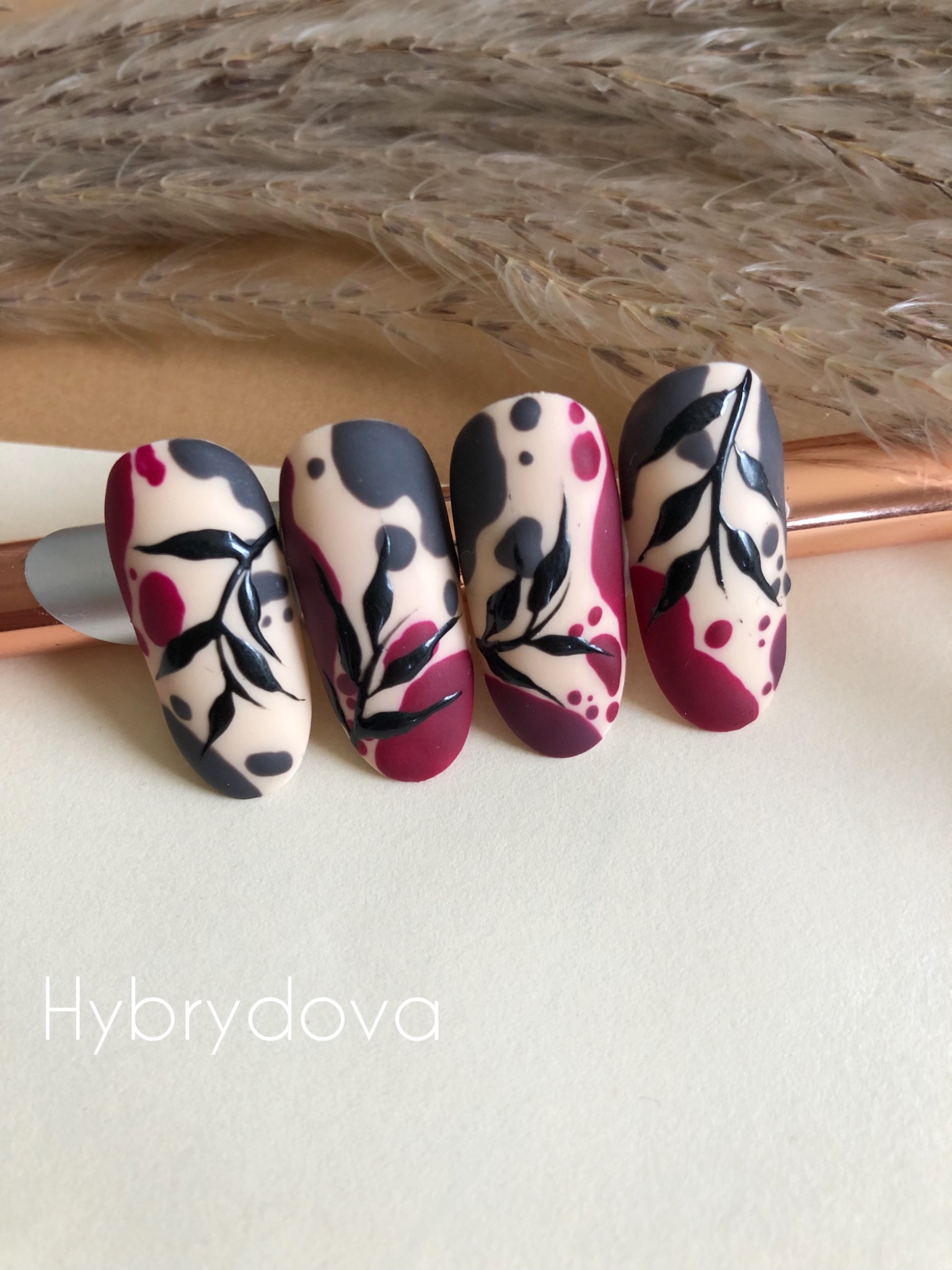 Cztery pomalowane paznokcie z abstrakcyjnym wzorem liści na beżowym tle, z elementami w kolorze burgundu i szarości.