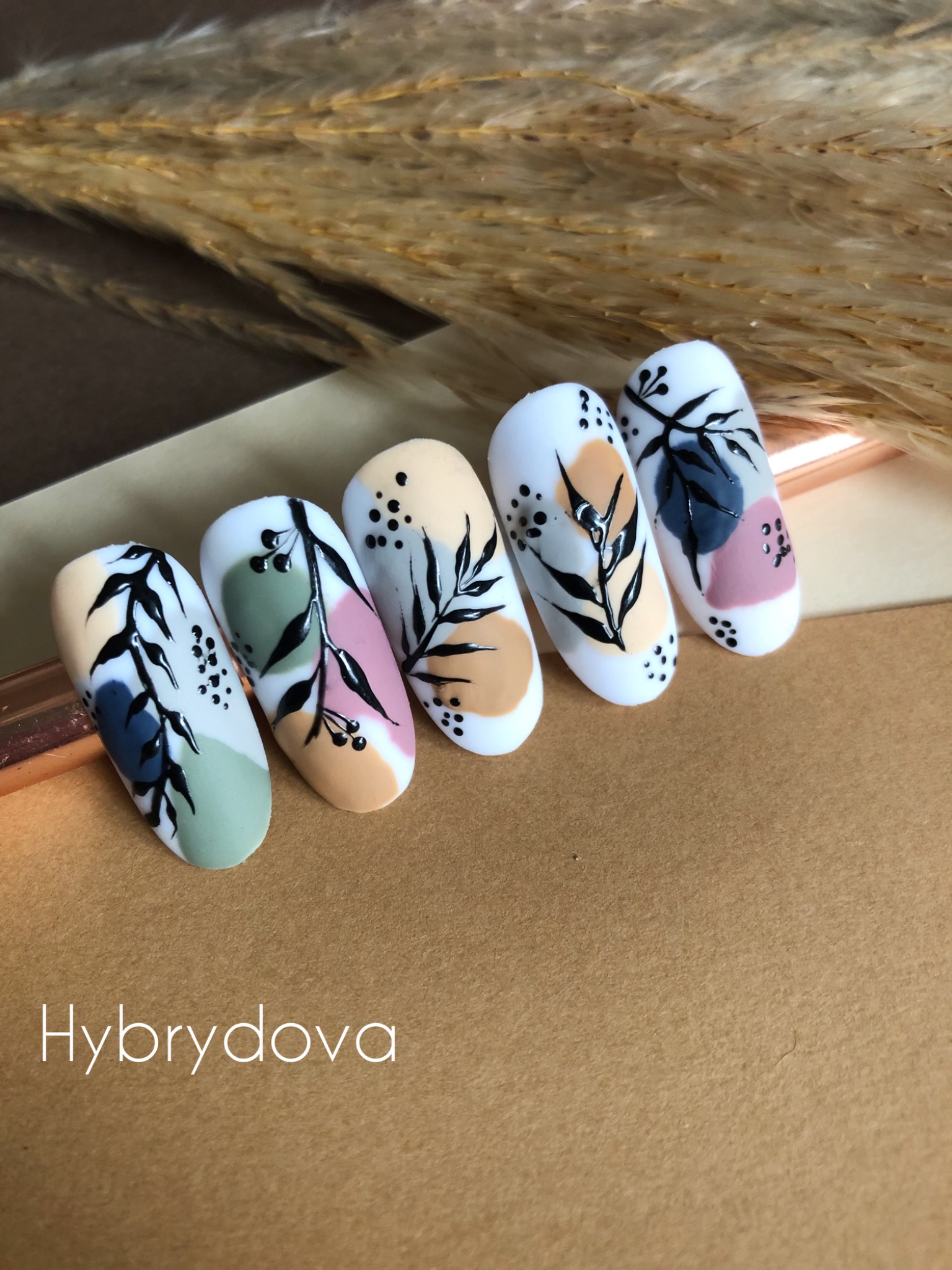 Pięć pomalowanych paznokci z abstrakcyjnym wzorem liści i kropek na białym tle, ułożonych na brązowym tle z dekoracyjną trawą w tle, prezentujących artystyczne zdobienie.
