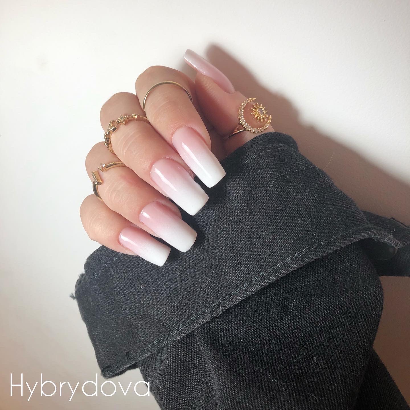 Dłoń z długimi, kwadratowymi paznokciami w stylu ombre (różowo-białe przejście), ozdobiona złotymi pierścionkami, na tle czarnej kurtki jeansowej i białej ściany.