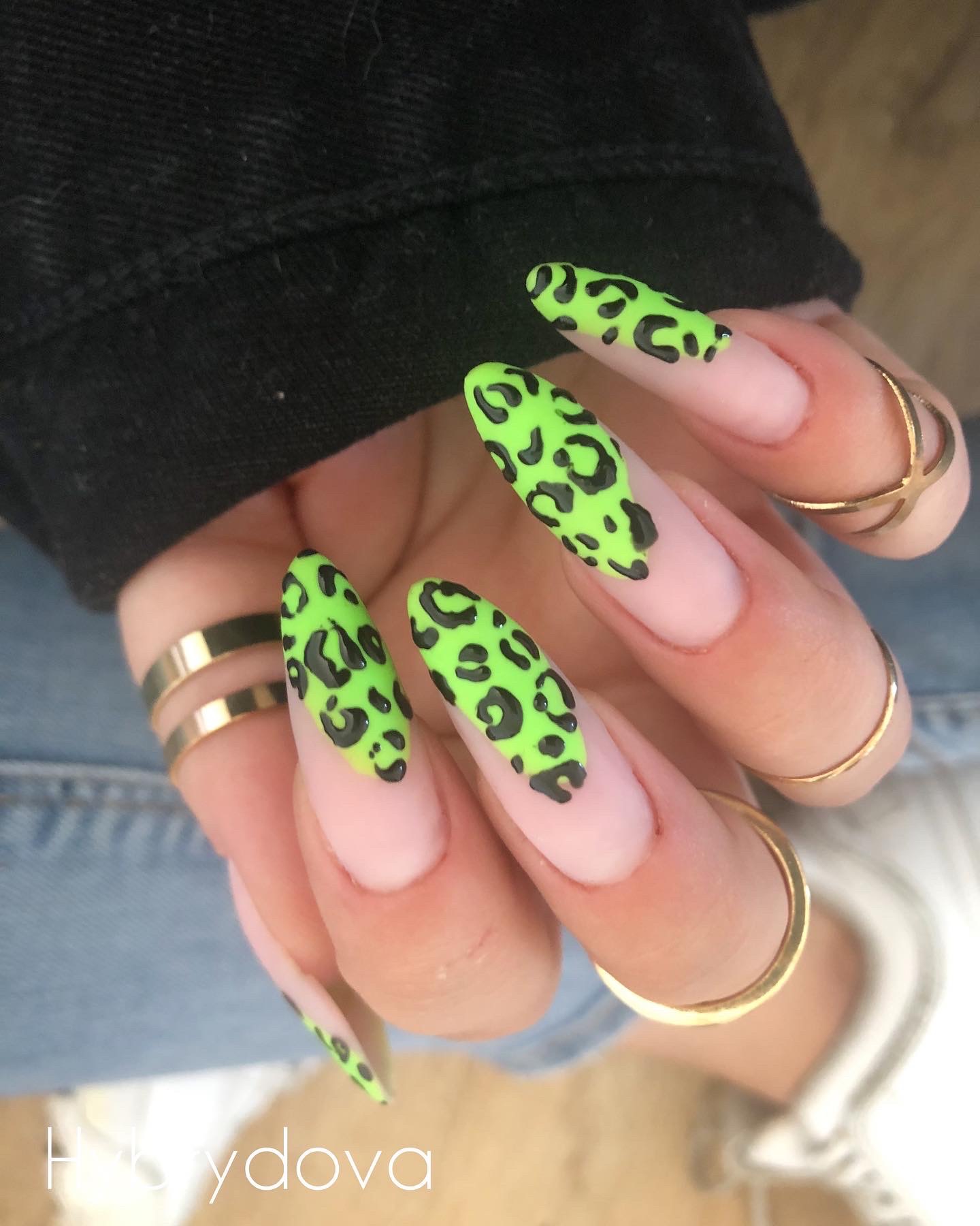 Dłoń z długimi paznokciami w kształcie migdała, pomalowanymi na jasny róż z neonowo-zielonym, panterkowym wzorem. Na palcach złote pierścionki.