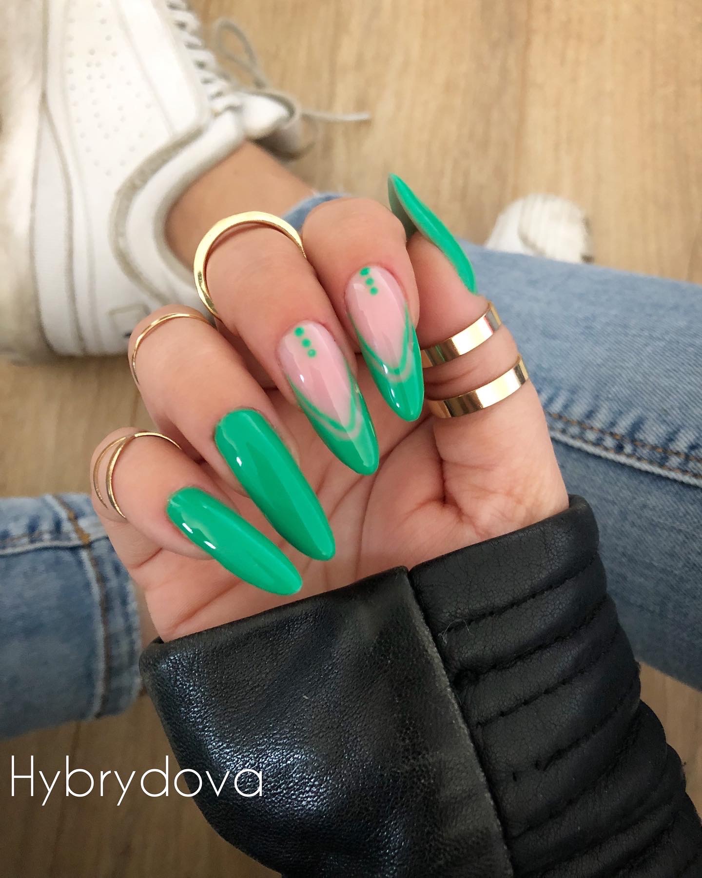 Dłoń z długimi, błyszczącymi, zielonymi paznokciami; jeden paznokieć z różowym tłem i zielonym geometrycznym wzorem oraz trzema zielonymi kropkami; złote pierścionki na palcach, skórzana kurtka...