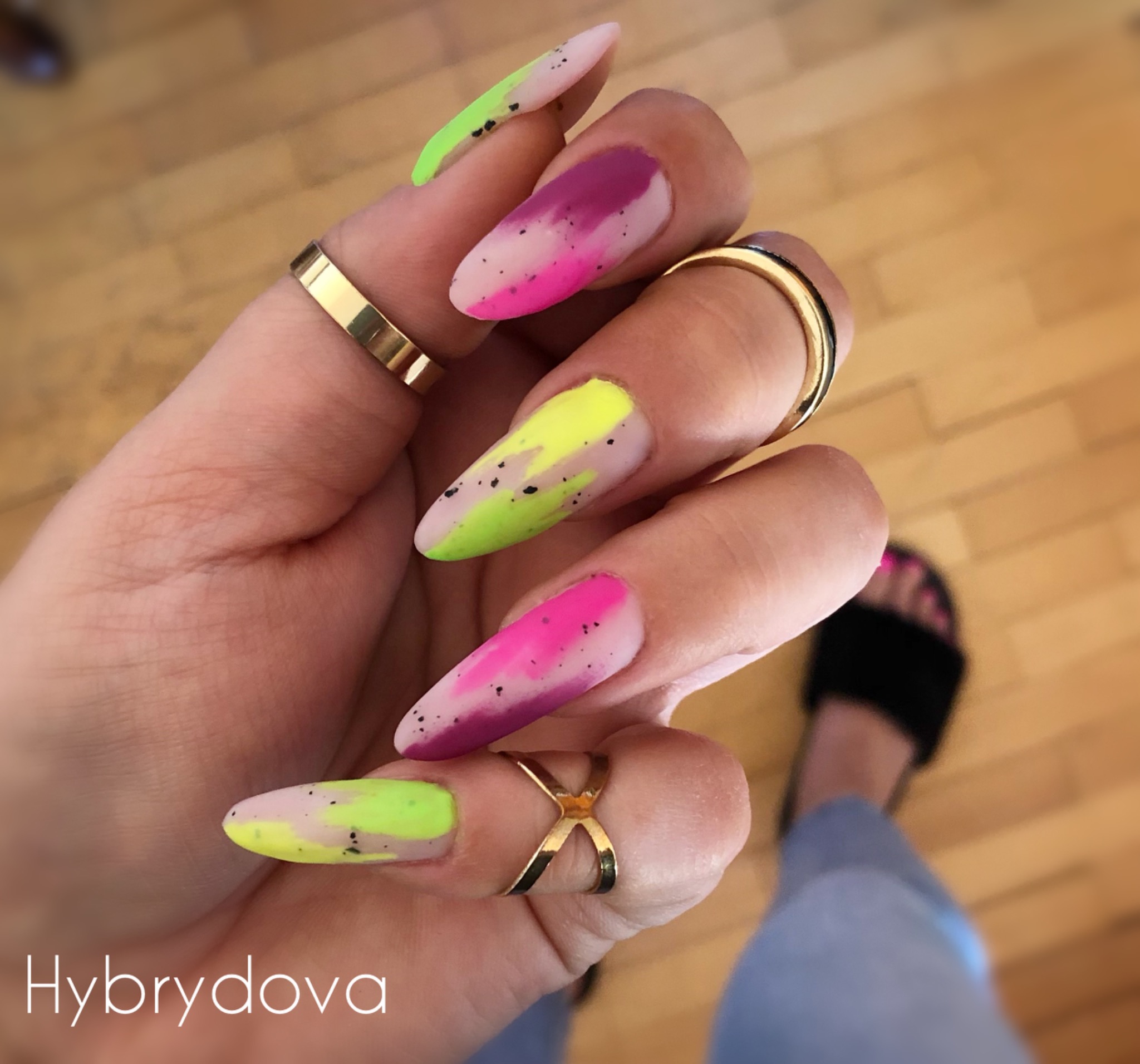 Dłoń z pomalowanymi paznokciami w odcieniach neonowej zieleni, różu i fioletu, zdobionymi czarnymi kropkami, z kilkoma złotymi pierścionkami na palcach, na tle drewnianej podłogi.