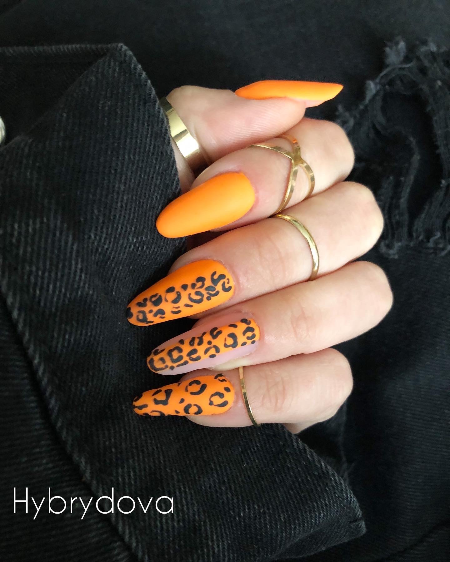 Dłoń z pomarańczowymi paznokciami w kształcie migdała, ozdobionymi czarnym wzorem panterki, nosząca złote pierścionki, na tle czarnej kurtki jeansowej.