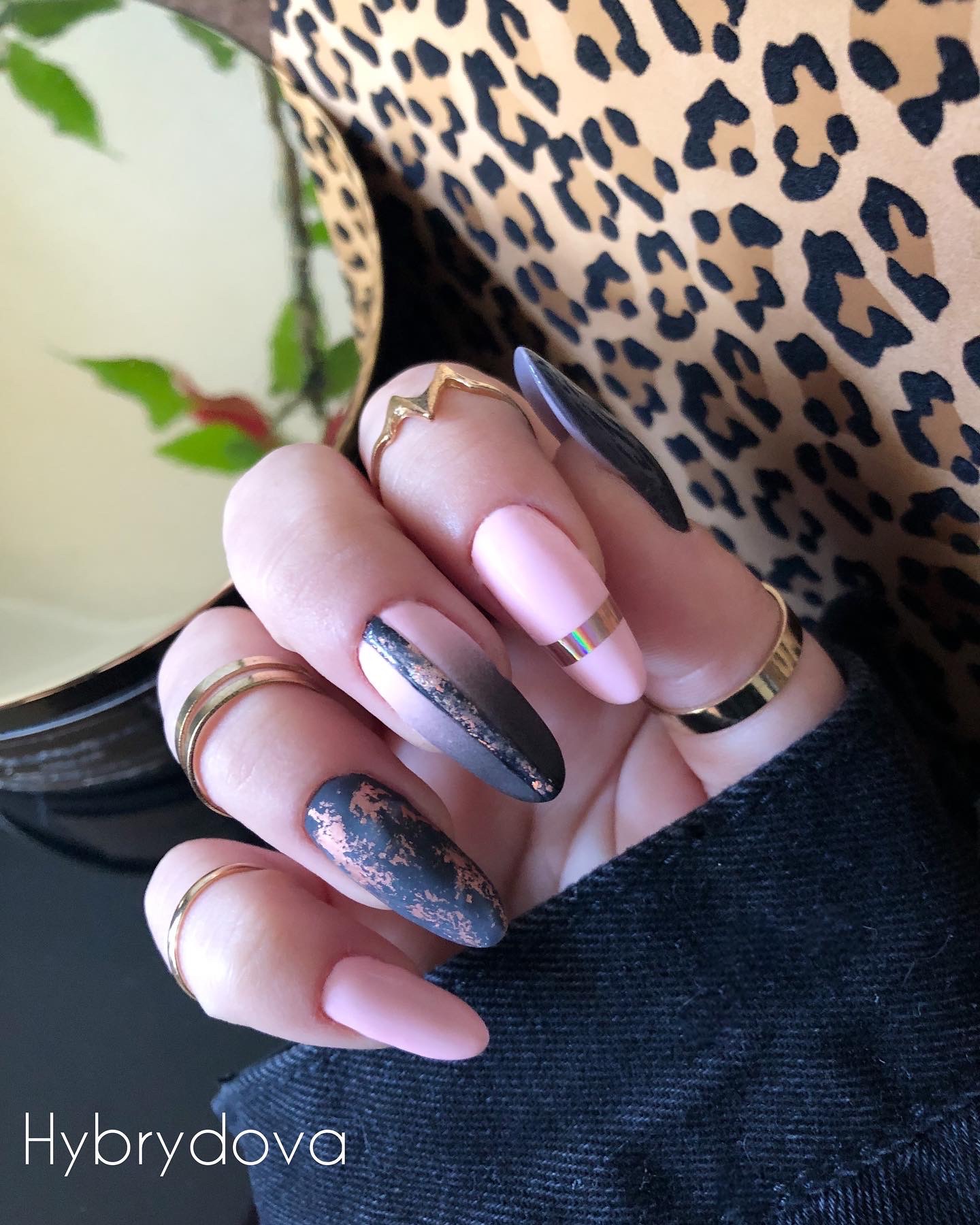 Dłoń z pomalowanymi na różowo i czarno paznokciami w kształcie migdała, ozdobionymi złotą folią i złotymi pierścionkami, na tle w panterkę i czarnej jeansowej kurtki.