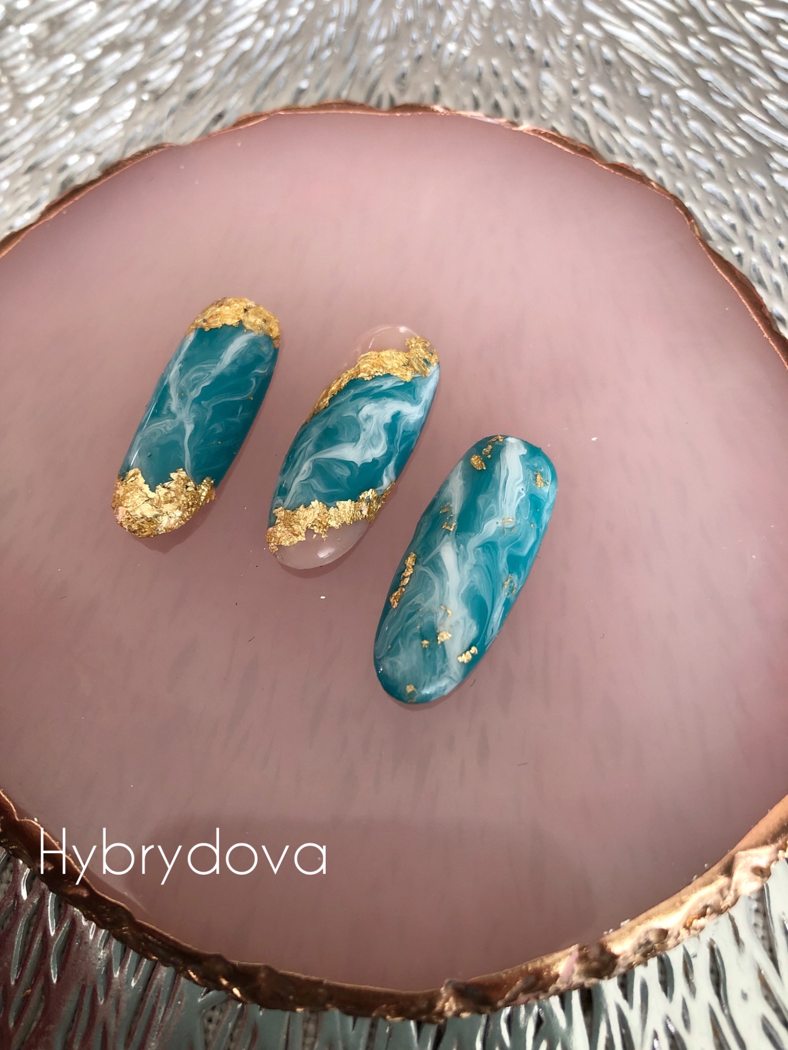 Trzy sztuczne paznokcie z turkusowo-białym marmurkowym wzorem i złotymi płatkami nałożone na różową, okrągłą podstawkę.
