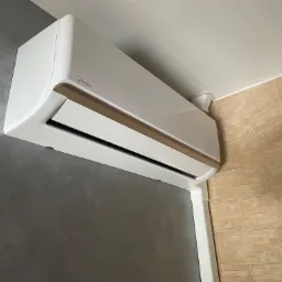 Biała klimatyzacja marki Midea zamontowana na ścianie w pomieszczeniu z szarą ścianą i beżowymi płytkami, widoczny plastikowy kanał instalacyjny.