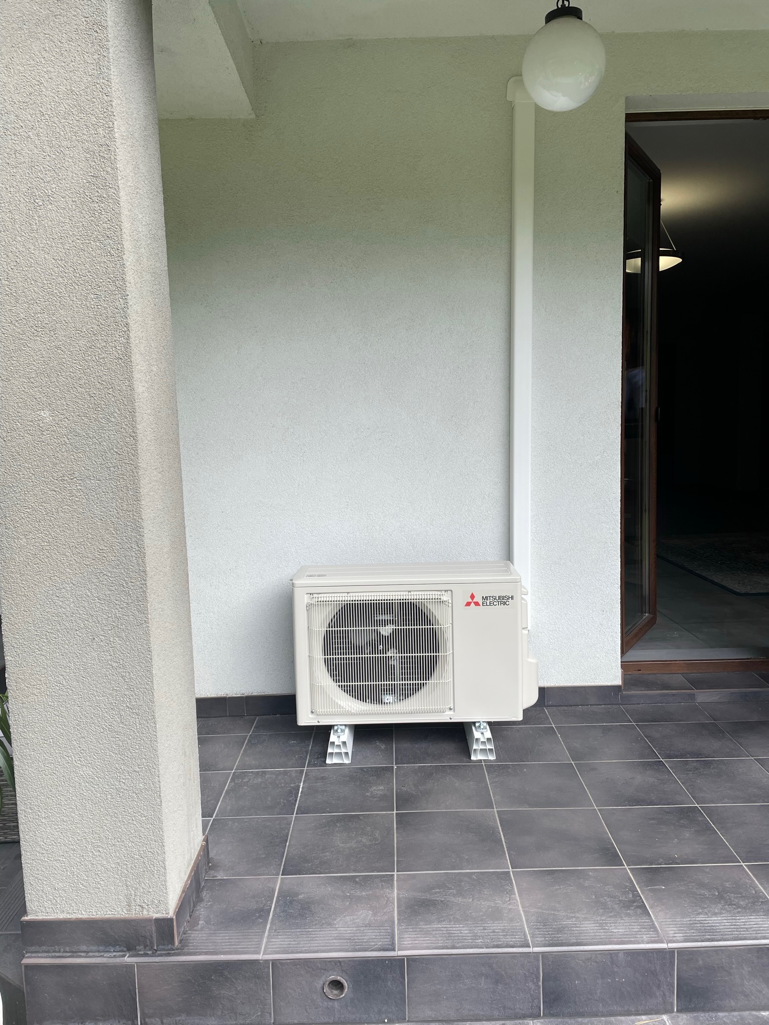 Zewnętrzna jednostka klimatyzacji Mitsubishi Electric zamontowana na szarych płytkach tarasowych przy ścianie budynku z widoczną rurą odprowadzającą skropliny i oświetleniem zewnętrznym.