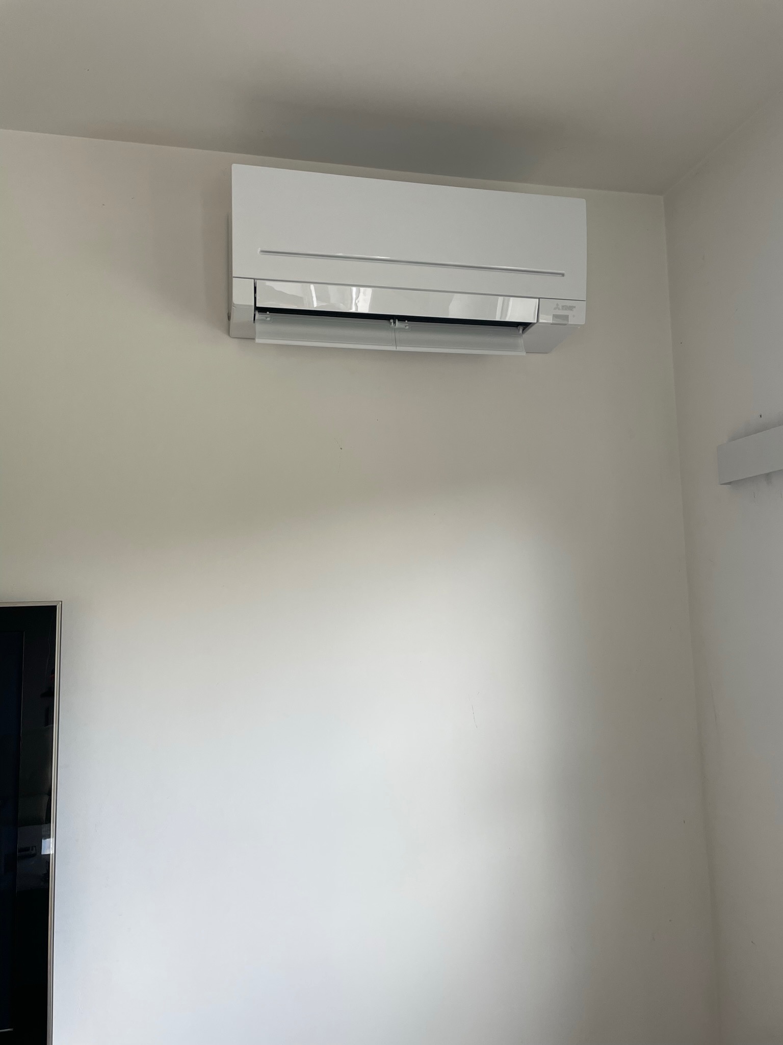 Biały klimatyzator Mitsubishi Electric zamontowany na jasnej ścianie w pomieszczeniu, widoczny fragment czarnej ramy telewizora po lewej stronie.