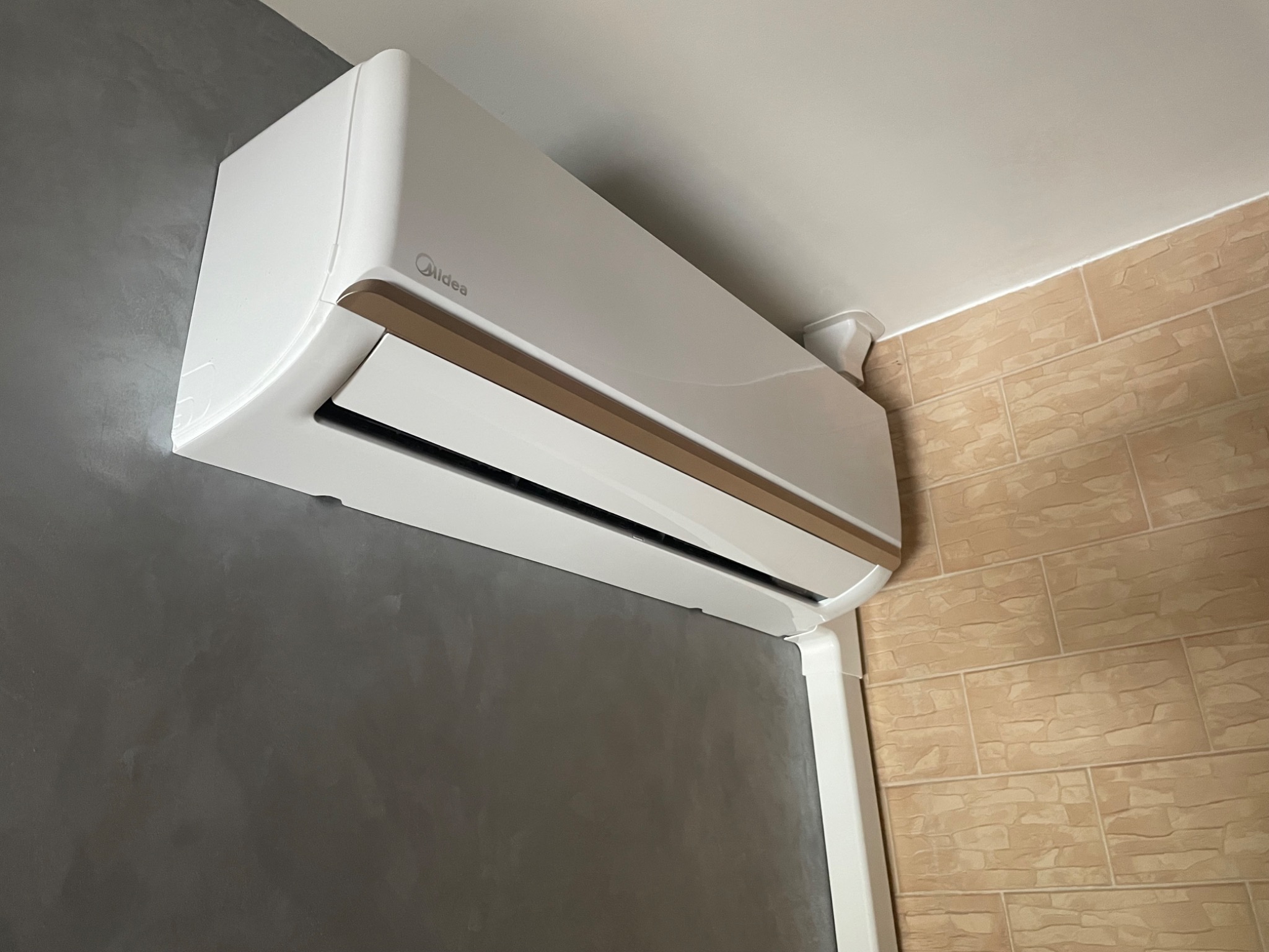 Biała klimatyzacja marki Midea zamontowana na ścianie w pomieszczeniu z szarą ścianą i beżowymi płytkami, widoczny plastikowy kanał instalacyjny.