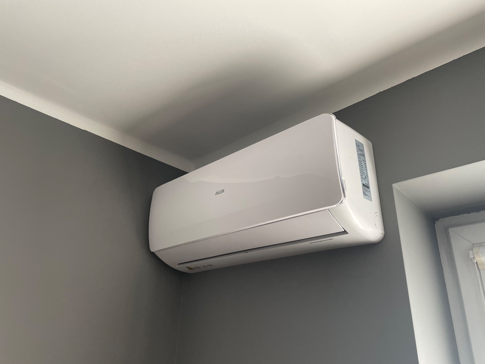 Biała jednostka klimatyzacji marki AUX zamontowana na szarej ścianie w rogu pokoju, widoczna z boku, obok okna.