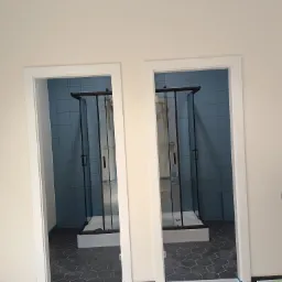 Dwie kabiny prysznicowe z czarnymi ramami, widoczne przez białe futryny drzwiowe, na podłodze heksagonalne płytki, ściany wyłożone niebieskimi płytkami.