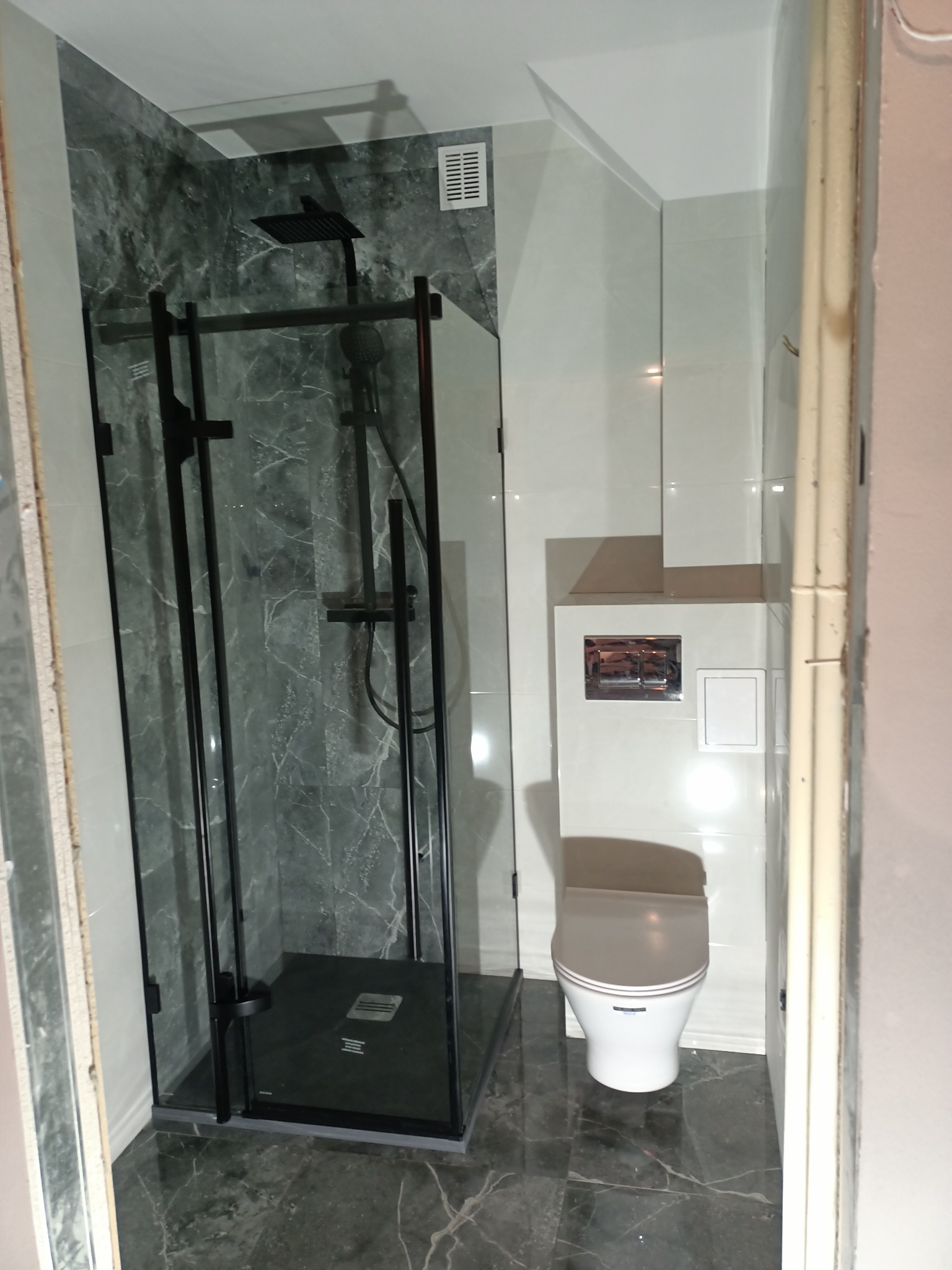 Nowoczesna łazienka z kabiną prysznicową typu walk-in z czarnymi profilami, marmurowymi płytkami i wiszącą toaletą z podtynkowym systemem spłukiwania.