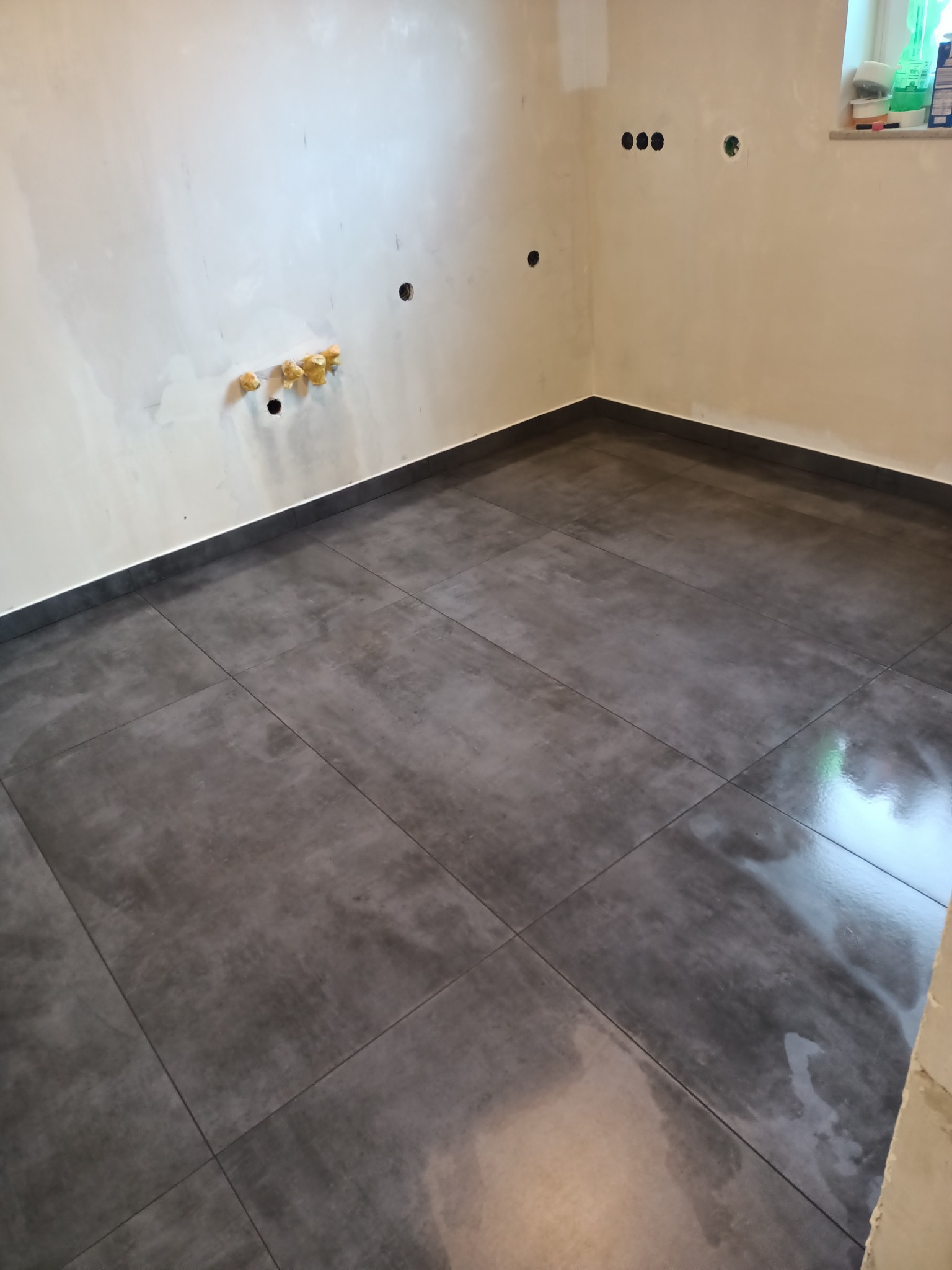 Wykończona podłoga z ciemnoszarych, matowych płytek w pomieszczeniu z surowymi, jasnymi ścianami przygotowanymi do malowania, widoczne otwory na gniazdka elektryczne zabezpieczone taśmą.