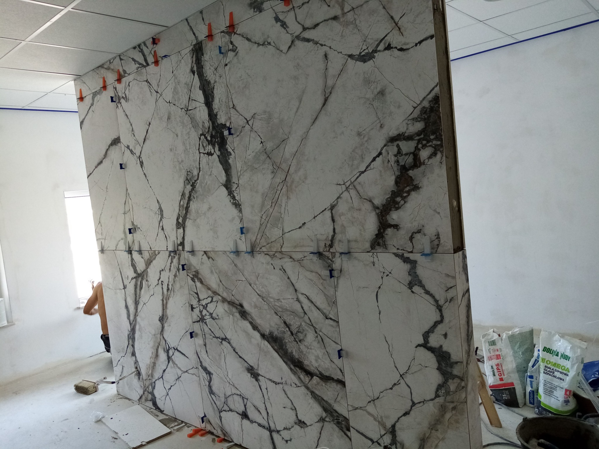 Montaż płyt imitujących marmur na ścianie, widoczne krzyżyki dystansowe, worki z zaprawą w tle, fragment sylwetki pracownika przy oknie.