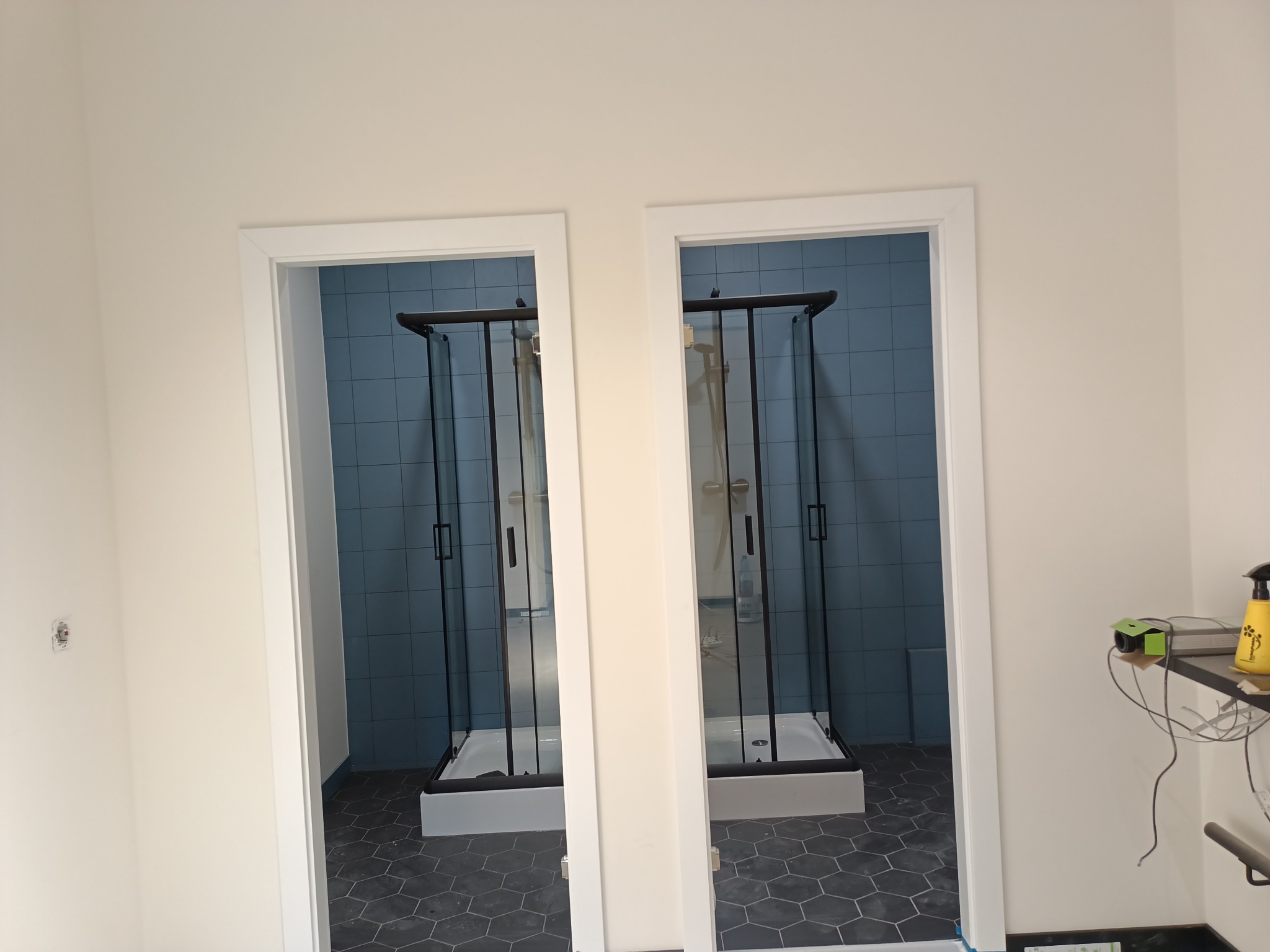Dwie kabiny prysznicowe z czarnymi ramami, widoczne przez białe futryny drzwiowe, na podłodze heksagonalne płytki, ściany wyłożone niebieskimi płytkami.