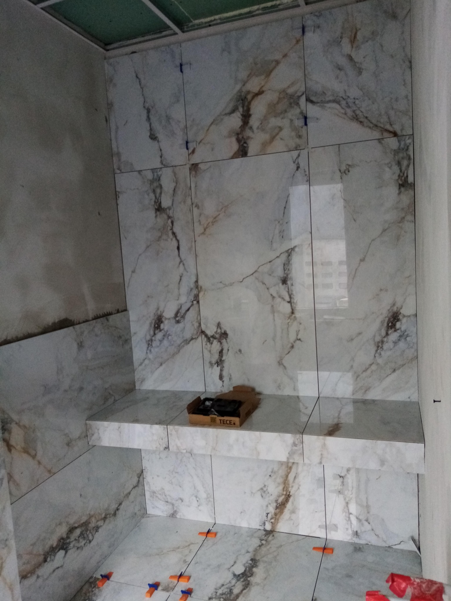 Łazienka w trakcie wykańczania, widoczne płytki imitujące marmur na ścianach i półce, plastikowe kliny do poziomowania płytek na podłodze, pudełko z elementami instalacyjnymi TECE.