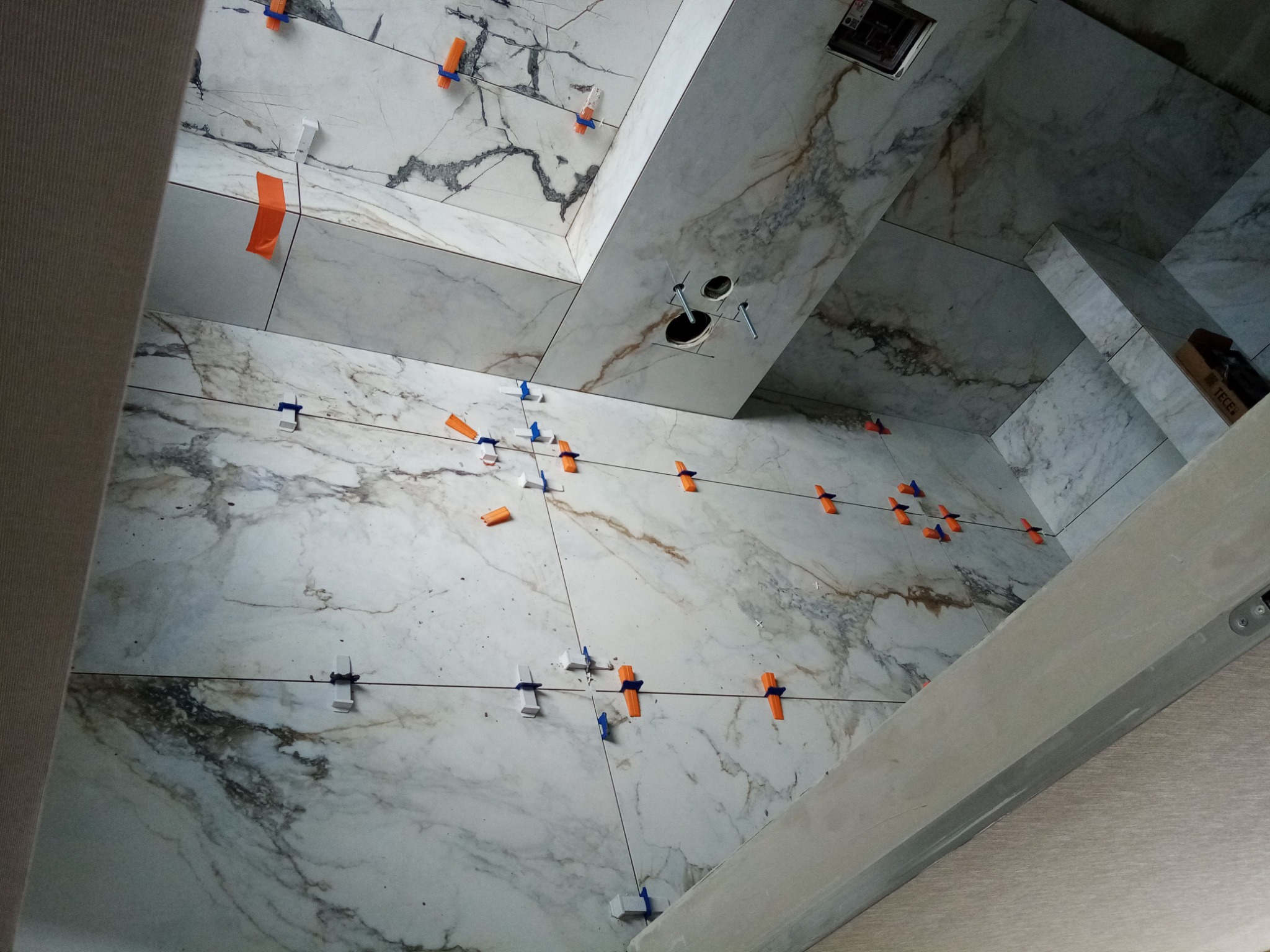 Układanie płytek imitujących marmur na ścianach łazienki w Szyk, z użyciem klipsów i poziomujących klinów, widoczne otwory na armaturę.