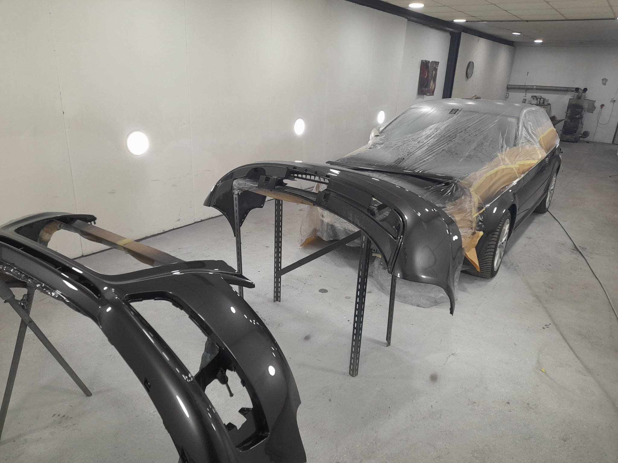 Przygotowany do lakierowania samochód owinięty folią ochronną, zdemontowane i polakierowane zderzaki na stojakach w warsztacie.