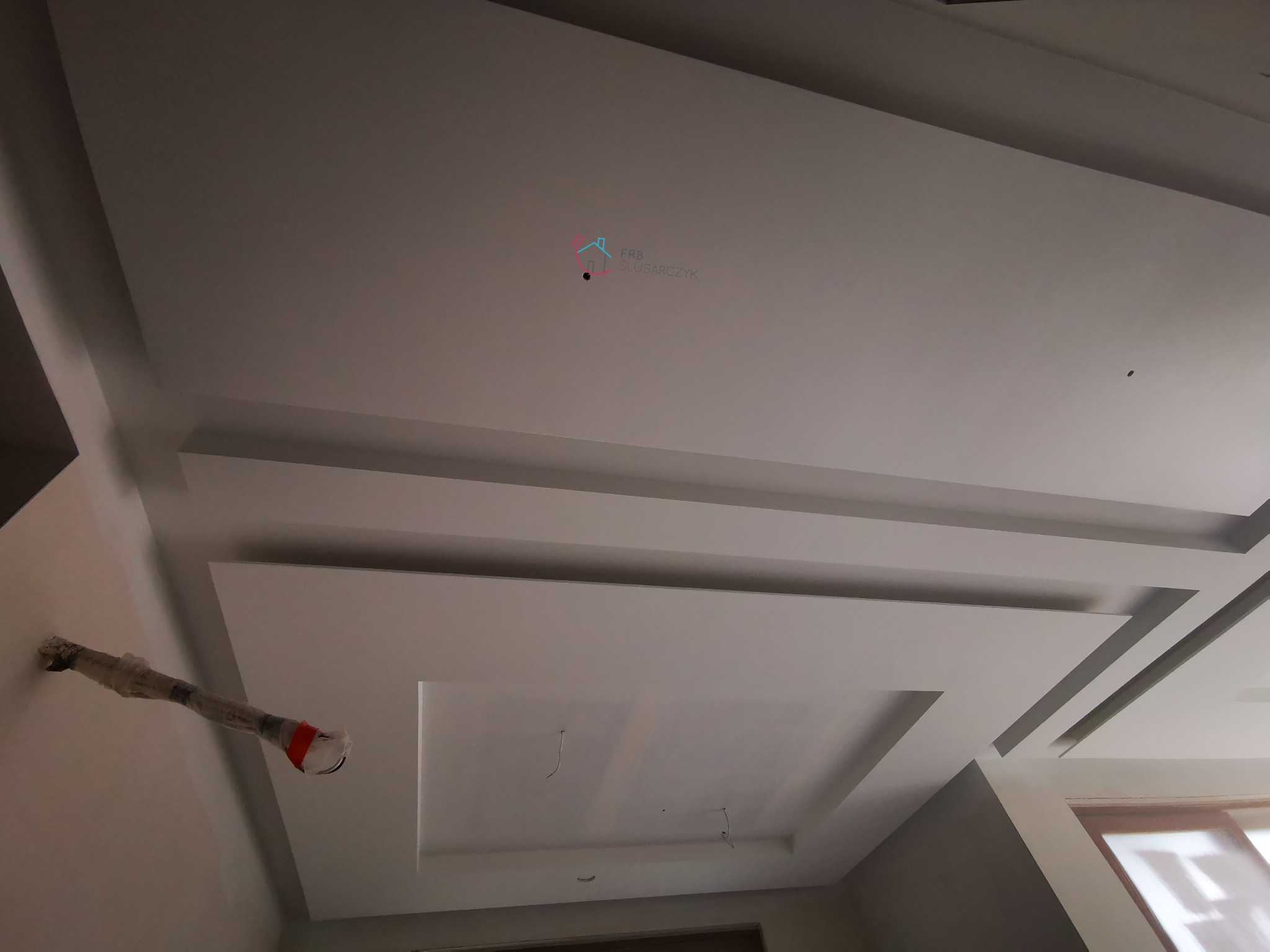 Sufit podwieszany z gładzi w trakcie wykańczania, widoczne kable i zabezpieczona lampa, logo firmy na suficie.