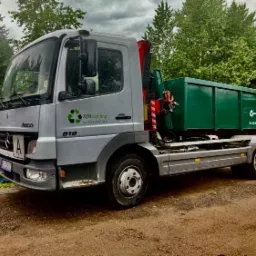 Srebrna ciężarówka Mercedes Atego 918 z zielonym kontenerem na gruz, z widocznym logo firmy AJRecykling i numerem telefonu, stojąca na ziemi w pochmurny dzień.