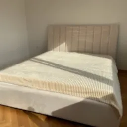 Beżowe łóżko tapicerowane z wysokim, pionowo pikowanym zagłówkiem, przykryte kremowym pledem w prążki, na parkiecie w jasnym, minimalistycznym wnętrzu.