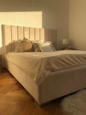 Beżowe łóżko tapicerowane z pionowymi przeszyciami na wezgłowiu, oświetlone popołudniowym słońcem, z dekoracyjnymi poduszkami i pledem w pasy, na parkiecie w jodełkę.