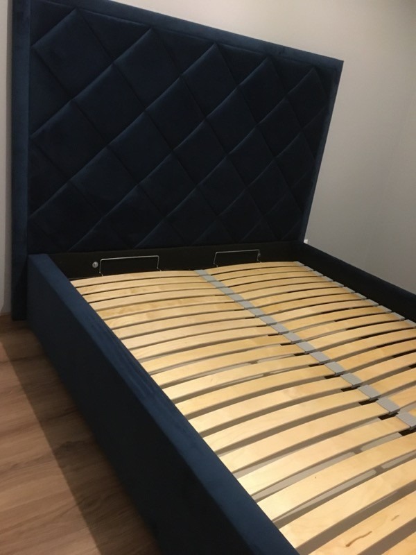 Niebieskie łóżko tapicerowane z pikowanym zagłówkiem w romby i drewnianym stelażem listwowym, widok z boku.