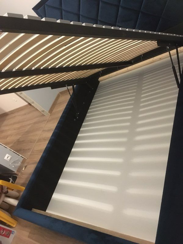 Otwarta rama łóżka tapicerowanego z niebieskim welurem i drewnianymi listwami stelaża, widoczne mechanizmy podnoszące, wnętrze białe, ujęcie z dołu pod kątem.