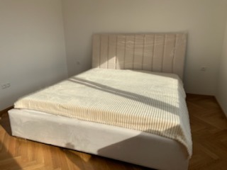 Beżowe łóżko tapicerowane z wysokim, pionowo pikowanym zagłówkiem, przykryte kremowym pledem w prążki, na parkiecie w jasnym, minimalistycznym wnętrzu.