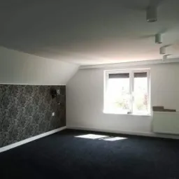Poddasze po renowacji w Czarnkowie z tapetą w liście, nowym dywanem i świeżo pomalowanymi ścianami, widoczne lampy sufitowe i okno.