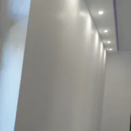 Świeżo pomalowane na biało ściany z widocznym połyskiem, oświetlone rzędem wbudowanych punktowych lamp sufitowych, z fioletowym akcentem na krawędzi sufitu.