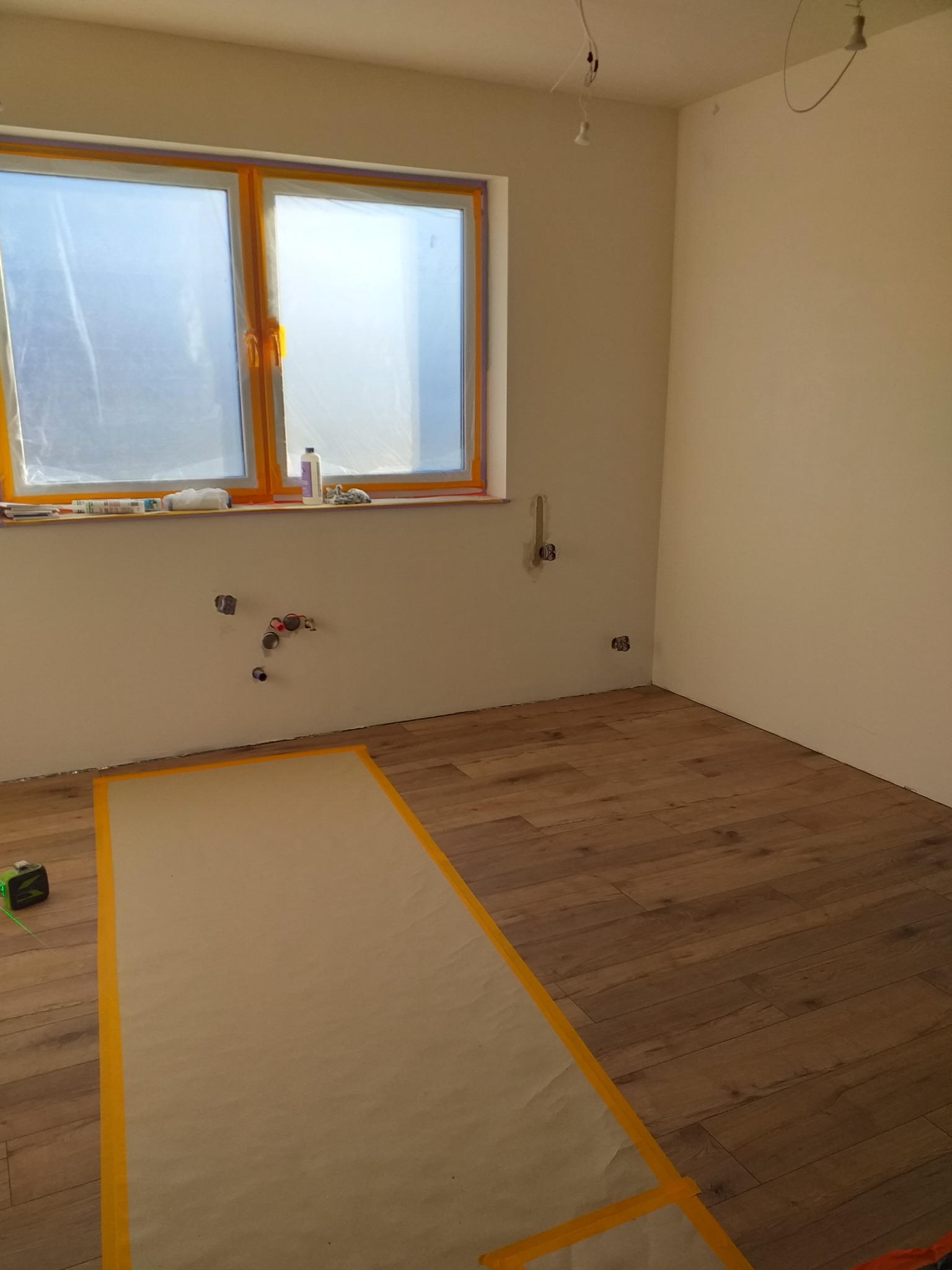 Pokój w trakcie remontu: Częściowo ułożone panele podłogowe z zabezpieczoną folią powierzchnią, okno zaklejone folią ochronną, widoczne instalacje na ścianie.