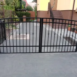 Czarna, metalowa brama wjazdowa z dekoracyjnymi elementami, zamontowana na posesji z kostką brukową i budynkiem w tle.