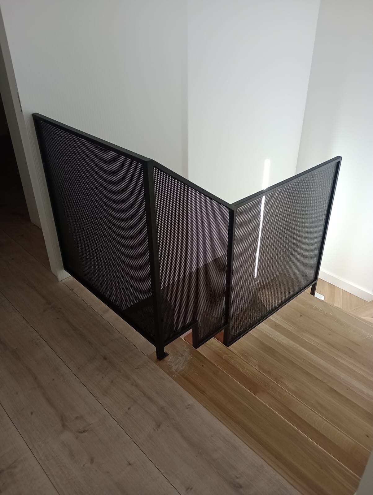 Nowoczesna, czarna, metalowa balustrada schodowa z perforowanej blachy, zamontowana na drewnianych schodach w jasnym wnętrzu.