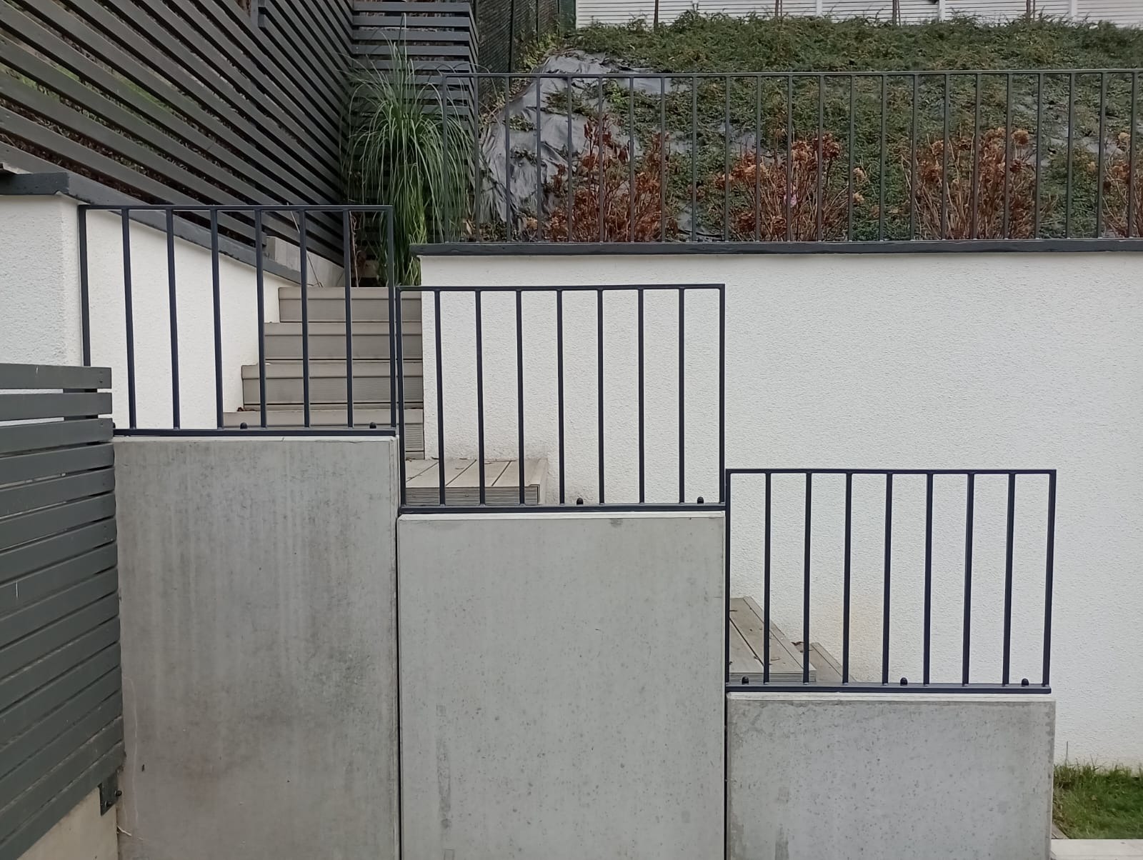 Schody zewnętrzne z betonowymi podestami i czarną, minimalistyczną balustradą z pionowych prętów, na tle białej ściany i zieleni na górze.
