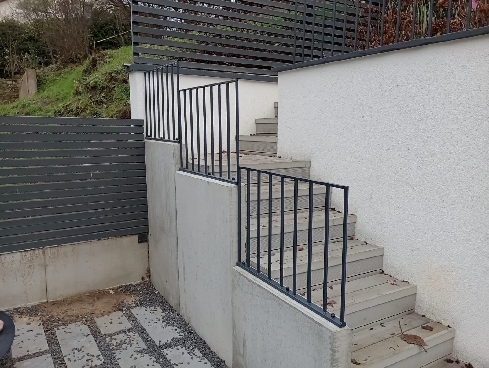 Metalowa balustrada schodowa na zewnątrz budynku, dopasowana kolorystycznie do poziomego ogrodzenia panelowego w tle, betonowe ściany oporowe i schody z drewnopodobnych desek.