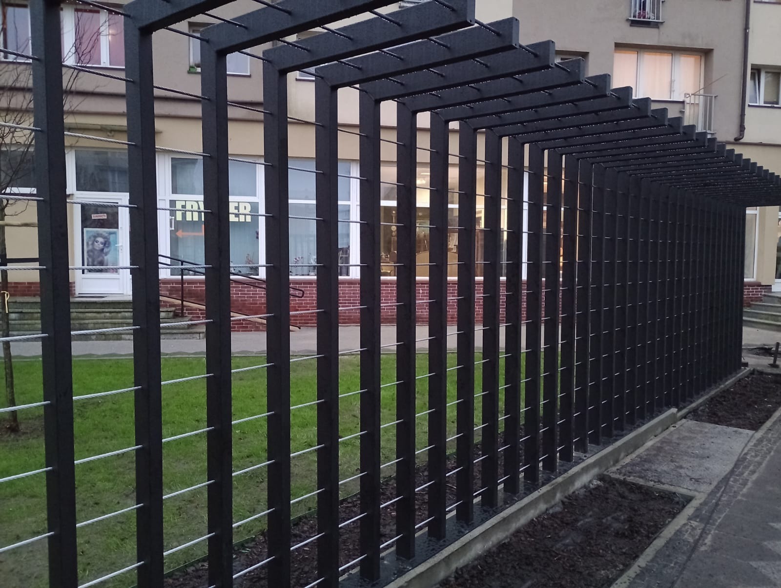 Nowoczesne, czarne ogrodzenie metalowe z poziomymi linkami stalowymi, widok z boku, na tle budynku z ceglanym cokołem i zielonym trawnikiem.