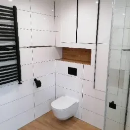 Nowoczesna łazienka z białymi, kwadratowymi płytkami, drewnianą podłogą i podwieszaną toaletą, widoczny grzejnik drabinkowy w kolorze czarnym oraz częściowo przeszklona kabina prysznicowa.