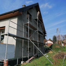 DAMTES USŁUGI WIELOBRANŻOWE DAMIAN DROBNY - Dom w trakcie ocieplania elewacji, widoczne rusztowania, częściowo ocieplona ściana z szarym tynkiem, błękitne niebo w tle. Ujęcie z dołu, z prawej strony budynku.