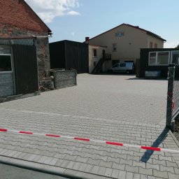 DAMTES USŁUGI WIELOBRANŻOWE DAMIAN DROBNY - Szara kostka brukowa ułożona na posesji, częściowo odgrodzona taśmą ostrzegawczą. W tle budynki i zaparkowany samochód dostawczy.
